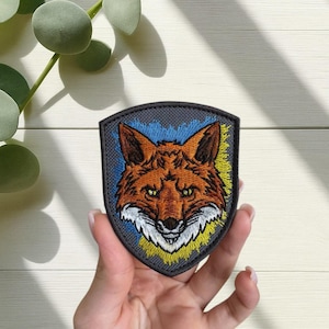 May include: Embroidered patch featuring a detailed fox head with orange fur, white muzzle, and yellow eyes. The fox is set against a shield-shaped backdrop with blue and yellow accents. The patch is gray with a textured appearance.
