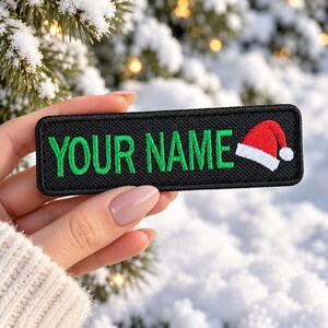 May include: A rectangular black embroidered patch featuring the green text YOUR NAME and a small red and white Santa hat icon on the right side. The patch has a black stitched border and a textured fabric surface.