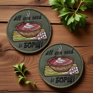 Pode incluir: Dois patches redondos bordados com o texto "All you need is БОРЩ!" acima de uma tigela de borscht. Os patches são verde-oliva com uma tigela de sopa marrom, creme branco e cebolinha verde. O fundo é uma superfície de madeira.