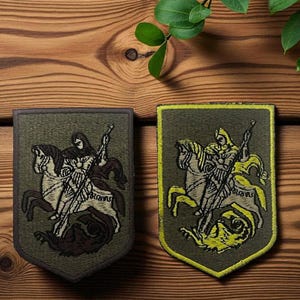 Pode incluir: Dois patches bordados em forma de escudo com um cavaleiro a cavalo matando um dragão. Os patches têm um fundo verde. Um tem uma borda marrom, o outro uma borda amarela. O cavaleiro e o cavalo são em tons de marrom e branco.