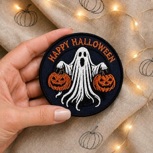 May include: A circular embroidered patch with a dark blue background featuring a white ghost holding two orange jack-o'-lanterns. The text "HAPPY HALLOWEEN" is stitched in orange thread in an arc along the top edge of the circular patch.