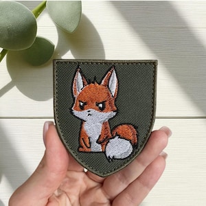 May include: Embroidered patch with a cartoon fox in orange and white, on a green shield. The fox has a grumpy expression. The patch is designed for application on clothing or accessories.