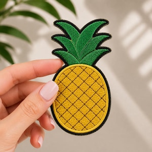 Puede incluir: Un parche bordado con forma de piña que presenta un cuerpo de fruta amarillo con una textura de cuadrícula y una parte superior de hojas verdes. Todo el diseño está delineado con un borde negro grueso, creando una forma clara para este accesorio.