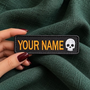 Puede incluir: Un parche rectangular bordado en negro con el texto YOUR NAME en letras mayúsculas amarillas, seguido de un icono de calavera blanca en el lado derecho. El parche tiene un borde cosido en negro y una base de tela texturizada.