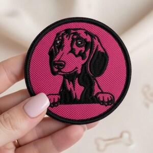 Pode incluir: Um emblema bordado circular com a ilustração de um cão dachshund preto sobre um fundo magenta vibrante. O emblema tem uma borda preta espessa e mostra o cão com as patas dianteiras apoiadas na borda inferior do círculo.