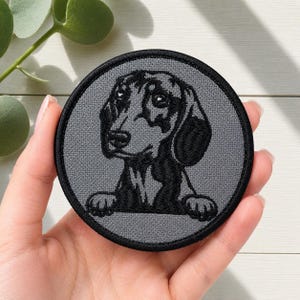 Peut inclure: Un écusson brodé circulaire représentant l'illustration d'un chien teckel en noir et gris. Le motif montre la tête et les pattes avant du chien sur un fond gris uni, entouré d'une bordure noire épaisse cousue sur le contour extérieur.
