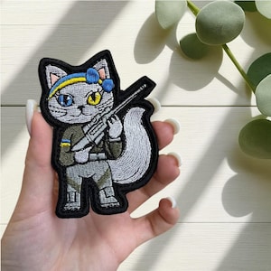 May include: Embroidered patch featuring a gray cat with blue and yellow accents, holding a rifle. The cat has one blue and one yellow eye, wearing a blue and yellow headband. The patch has a black border and is held in a hand.