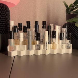 Può includere: Un vassoio esagonale bianco contiene una collezione di piccoli flaconi di profumo. I flaconi sono in vetro trasparente con tappi neri e contengono diverse fragranze. Il vassoio è su una superficie grigia.