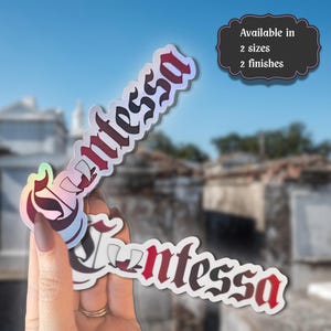 Könnte beinhalten: Zwei gestanzte Aufkleber mit dem Wort Contessa in einer gotischen Schriftart mit einem Farbverlauf von Rot zu Schwarz. Ein Aufkleber hat ein holografisches Finish, der andere ein mattes. Ein Textfeld zeigt Available in 2 sizes 2 finishes.