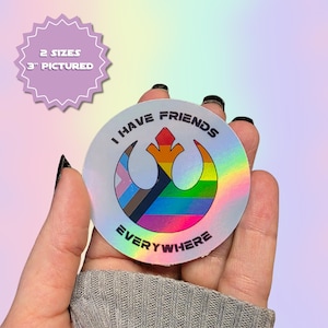 Può includere: Un adesivo olografico circolare con un simbolo stilizzato dell'Alleanza Ribelle riempito con un motivo a bandiera arcobaleno. Il testo "I HAVE FRIENDS EVERYWHERE" è stampato in lettere maiuscole nere attorno all'emblema centrale.