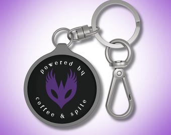 May include: Silver keychain with a round black and silver charm that says "powered by coffee & spite" with a purple winged mask in the center.