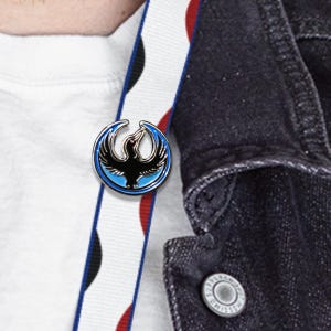 May include: A round, silver-colored pin with a black bird design, set against a blue background. The pin is attached to a white strap with red and black accents, and a dark denim jacket.