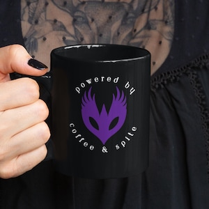 以下が含まれることがあります： 紫色の羽根のついたマスクのグラフィックと「powered by coffee & spite」というテキストが書かれた黒いコーヒーカップ。