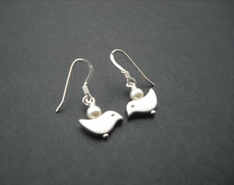 Puede incluir: Un par de pendientes de plata con una pequeña perla blanca y un diseño de pájaro.