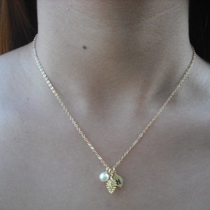 May include: A gold chain necklace with a small pine cone charm, a pearl, and a gold disc with the letter "H" on it.
