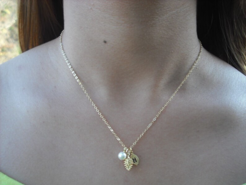 May include: A gold chain necklace with a small pine cone charm, a pearl, and a gold disc with the letter "H" on it.