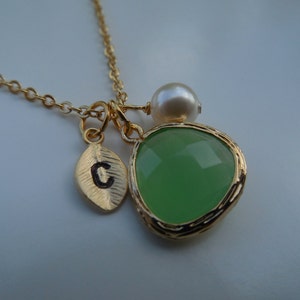 May include: A gold necklace with a green gemstone pendant, a small pearl charm, and a leaf charm with the letter "C" engraved on it.