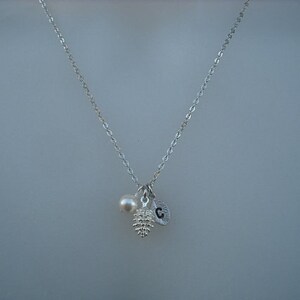 May include: A silver chain necklace with a small pearl, a silver pine cone charm, and a silver charm with the letter "C" hanging from it.