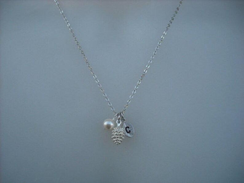 May include: A silver chain necklace with a small pearl, a silver pine cone charm, and a silver charm with the letter "C" hanging from it.