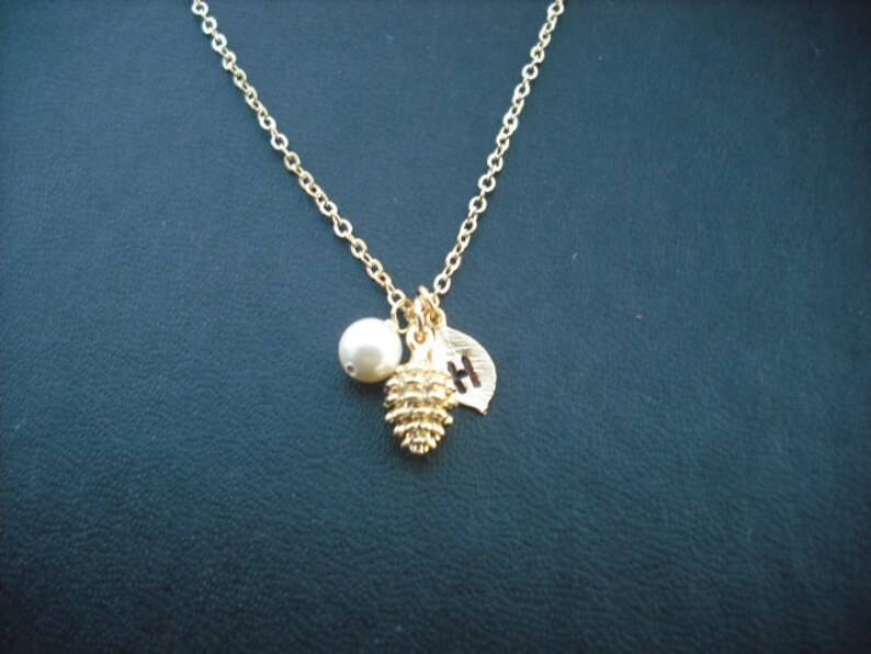 May include: A gold chain necklace with a small pearl, a gold pine cone charm, and a gold leaf charm with the letter "H" engraved on it.