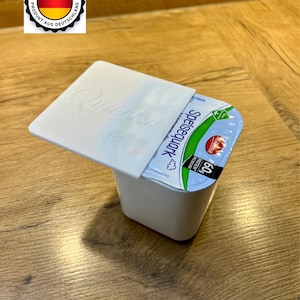 Peut inclure: Un récipient blanc de produit laitier Spiesequark avec le couvercle partiellement ouvert. Le récipient porte une étiquette bleue et blanche avec du texte et une feuille verte. Un autocollant "Made in Germany" est en haut à gauche.