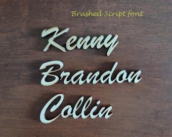 May include: Three wooden names, Kenny, Brandon, and Collin, are written in a brushed script font. The names are cut out of wood and have a light brown color.