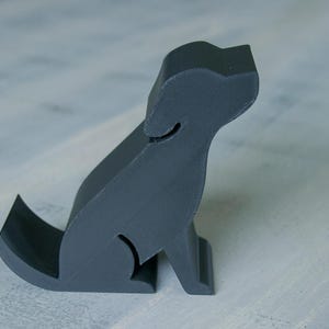 May include: A gray, 3D-printed dog figurine in a sitting position. The dog has a simple, stylized design with a curved tail and a cutout detail on the body. The figurine is on a light gray surface.