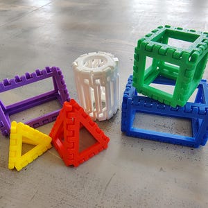 May include: A collection of 3D printed geometric shapes including a purple rectangular frame, a white cylindrical cage, a yellow triangle, a red pyramid, and two stacked blue and green cubes, all featuring a notched, interlocking edge design.