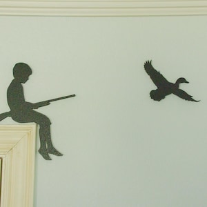 May include: A decorative wall art piece featuring a silhouette of a person with a rifle, seated on a frame corner, and a flying duck. The artwork is in a dark color, set against a light-colored wall, creating a hunting or wildlife theme.