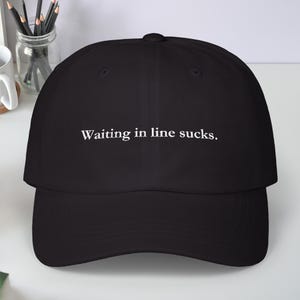 Puede incluir: Gorra de béisbol negra con el texto blanco "Waiting in line sucks." La gorra tiene una visera curva y una corona estructurada. El texto está en una fuente sans-serif.