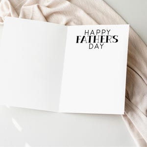 以下が含まれることがあります： 「HAPPY FATHERS DAY」の黒い文字が書かれた白いカード。カードは白い表面に置かれ、ベージュの布が周りに掛けられています。画像は明るく、柔らかい影があります。