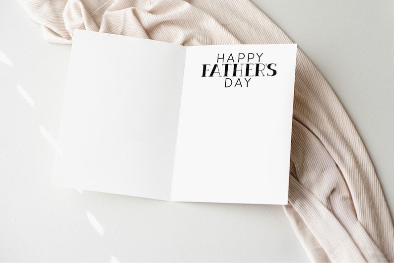 以下が含まれることがあります： 「HAPPY FATHERS DAY」の黒い文字が書かれた白いカード。カードは白い表面に置かれ、ベージュの布が周りに掛けられています。画像は明るく、柔らかい影があります。