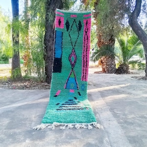 Peut inclure: Un tapis rectangulaire tissé à la main avec un fond turquoise et des motifs géométriques noirs, roses et bleus. Le tapis présente des formes de diamants et de triangles, avec une frange sur le bord inférieur. Le tapis est exposé à l'extérieur.