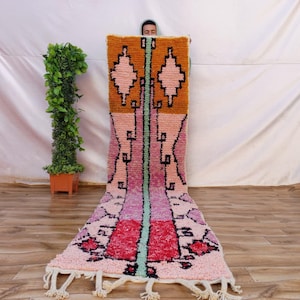 May include: A long, rectangular runner rug featuring a vibrant colour palette of mustard yellow, soft pink, magenta, and a central mint green vertical stripe. The rug displays geometric diamond patterns and black linear motifs with white fringe ends.