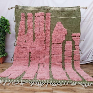 Peut inclure: Un tapis rectangulaire doté d'une base en vert olive texturé avec des bandes verticales en relief rose pelucheux de hauteurs et largeurs variées. Le tapis présente une bordure supérieure tissée, des franges nouées et des pompons latéraux.