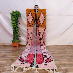 Op de afbeelding: Een kleurrijk, rechthoekig tapijt met een geometrisch ontwerp. Het tapijt heeft een combinatie van oranje, roze, rood en zwarte kleuren. Het tapijt heeft een franje aan de onderkant en wordt verticaal gepresenteerd.