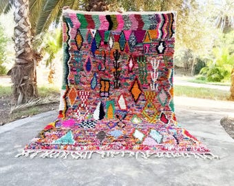Könnte beinhalten: Ein lebendiger, handgewebter Teppich mit farbenfrohem geometrischem Design. Der Teppich zeigt eine Mischung aus Formen, darunter Rauten, Dreiecke und Rechtecke, in Rosa, Grün, Orange und Blau. Der Teppich hat einen Fransenrand.