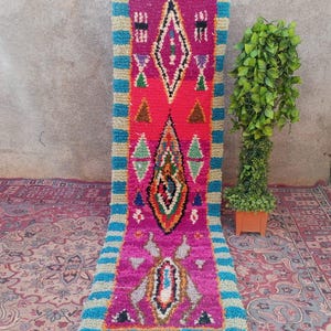 May include: A vibrant, rectangular Moroccan rug with a central design in pink and red, framed by turquoise stripes. The rug features geometric patterns in various colors and has a fringed edge. A small potted plant is visible to the right.