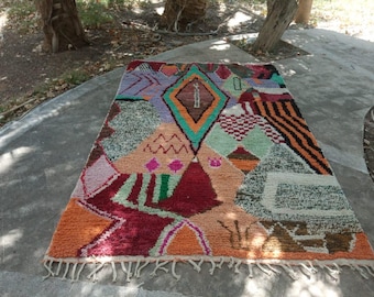 Könnte beinhalten: Ein farbenfroher Teppich mit geometrischen Mustern in verschiedenen Formen und Farben, darunter Orange, Rot, Grün und Lila. Der Teppich hat ein rautenförmiges Muster in der Mitte und einen Fransenrand. Ideal, um einen Raum aufzuhellen, ca. 180 cm x 120 cm.