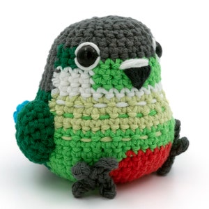 Könnte beinhalten: Ein gehäkeltes Papageienspielzeug aus grünem, weißem, grauem und rotem Garn. Der Papagei hat schwarze Augen und einen schwarzen Schnabel.