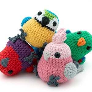 May include: Four brightly coloured, crocheted bird toys. The birds are in shades of yellow, green, red, and pink. Each bird has black eyes and detailed features, such as wings and beaks. The toys are handmade and appear soft and cuddly.