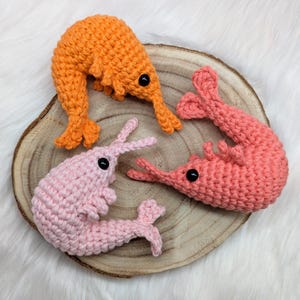 May include: Three crocheted shrimp toys in different colors: orange, pink, and coral. The shrimp are arranged on a wooden slice.