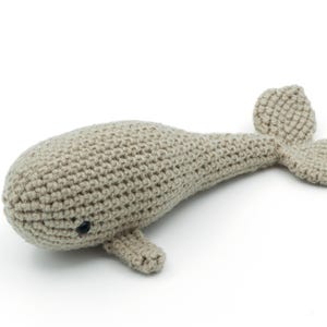 Puede incluir: Un juguete de peluche de ballena tejido a crochet en color beige claro. La ballena tiene un pequeño ojo negro y una cola larga y delgada.