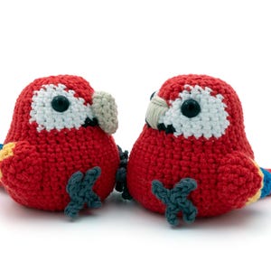 Puede incluir: Dos loros guacamayos rojos de crochet con ojos negros y blancos y detalles amarillos y azules. Los loros se miran.