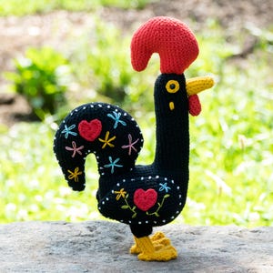 Puede incluir: Un gallo negro de ganchillo hecho a mano con cresta y barbilla rojas, pico y patas amarillas. El cuerpo del gallo está adornado con corazones bordados y diseños florales en varios colores. El gallo está sobre una superficie de piedra.
