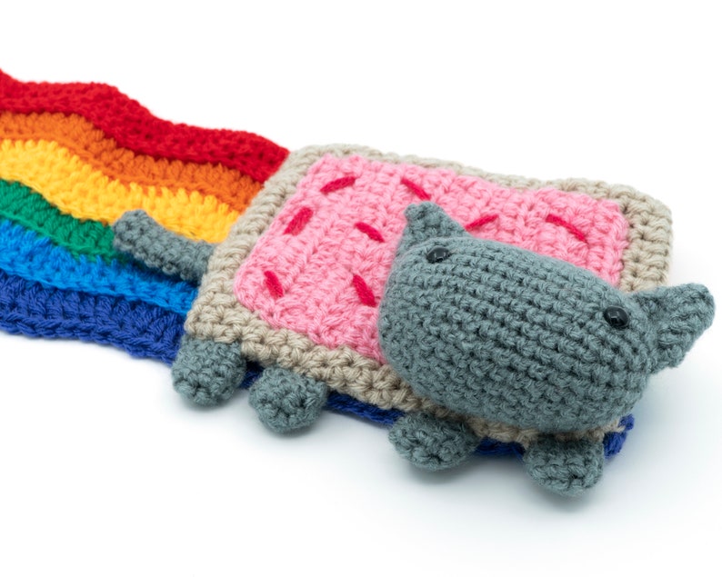 K&ouml;nnte beinhalten: Ein geh&auml;kelter Regenbogen mit einer grauen Katze, die ein geh&auml;keltes Toastkost&uuml;m in Rosa und Braun tr&auml;gt.