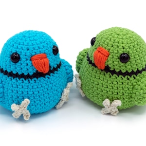 Könnte beinhalten: Zwei gehäkelte Vogel-Plüschtiere, eines blau und eines grün, mit schwarzen Augen und orangefarbenen Schnäbeln. Die Plüschtiere haben weiße gehäkelte Füße.