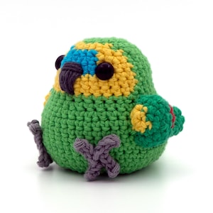 May include: A small, round, crocheted bird plushie featuring a vibrant green body, a yellow face with bright blue accents around the eyes, a dark purple beak, and dark purple feet. The bird has two large, round, black plastic safety eyes.