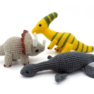 May include: Three crocheted dinosaur toys: a grey triceratops, a yellow and green brachiosaurus, and a grey plesiosaur.