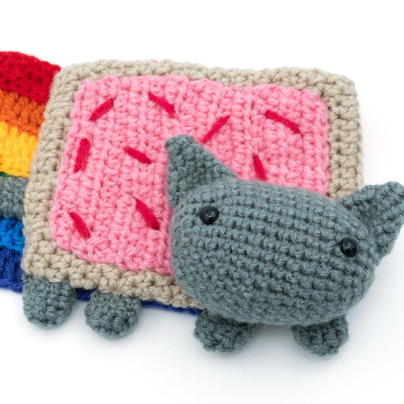 K&ouml;nnte beinhalten: Ein geh&auml;keltes Stofftierkatze, das ein rosa-braunes 'Nyan Cat'-Kost&uuml;m tr&auml;gt. Die Katze hat graues Fell, schwarze Augen und eine rosa Nase. Das Kost&uuml;m ist ein Quadrat mit einer rosa F&uuml;llung und einem braunen Rand. Die Katze sitzt auf einem wei&szlig;en Hintergrund.